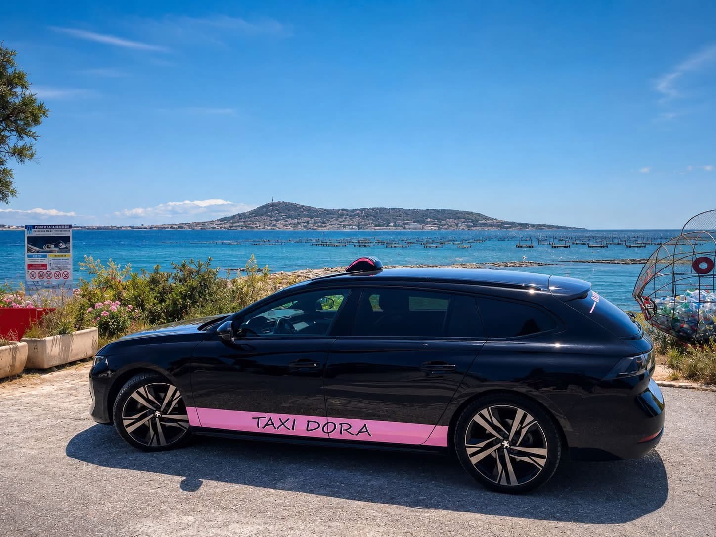 Peugeot noire de Dora avec une bande rose portant l’inscription Taxi Dora, stationnée au bord de l’eau.